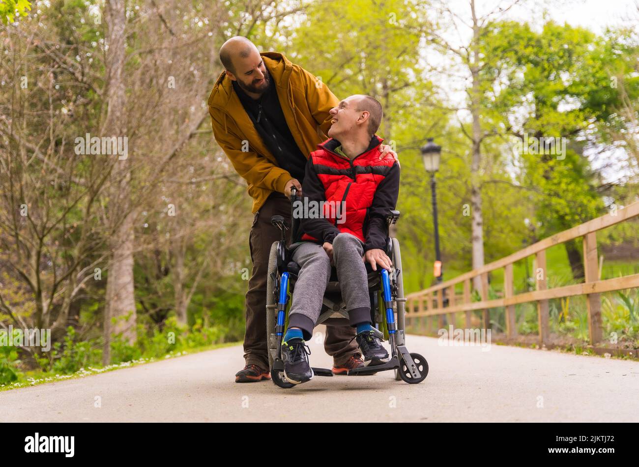 Paralyzed young man in the wheelchair being pushed by a friend in a public city park, strolling along a path having fun and smiling Stock Photo