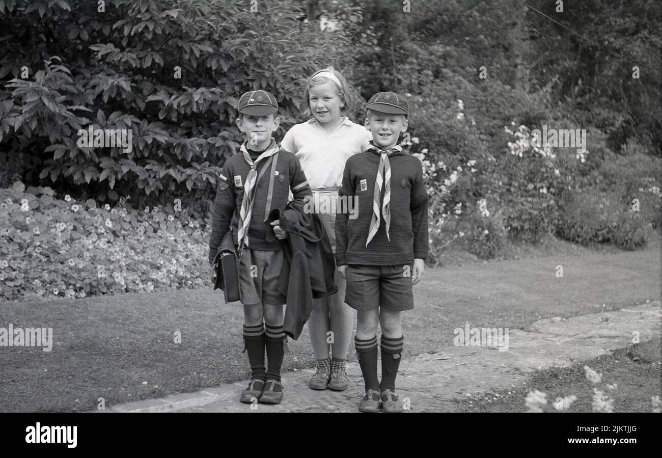1960s, historical, outside on a garden path, a girl standing for a photo with her two younger brothers, both wearing their Wolf cub scout uniform of the era, England, UK. Known as Wolf cubs a name originally derived from the Kipling story, The Jungle Book later in this era, young boy scouts began to become known simply as Cubs or Cub scouts, while retaining the traditional Wolf cub ceremonies such as the Grand Howl. Stock Photo