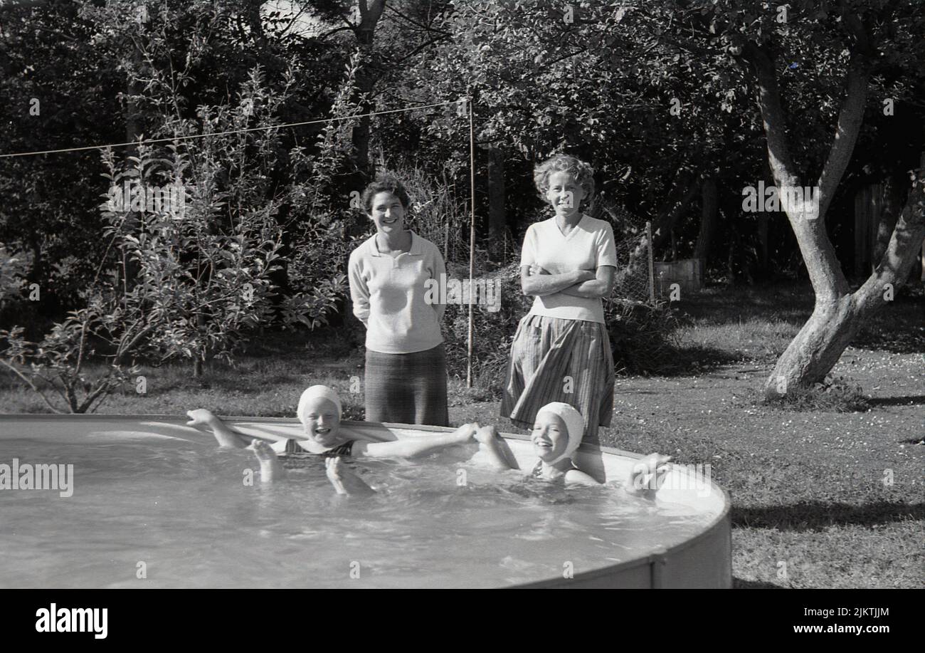 1963, historical, summertime and outside in a garden, two mothers standing with their young daughters, who are enjoying themselves lying in a large above ground swimming pool or tub, England, UK. Stock Photo