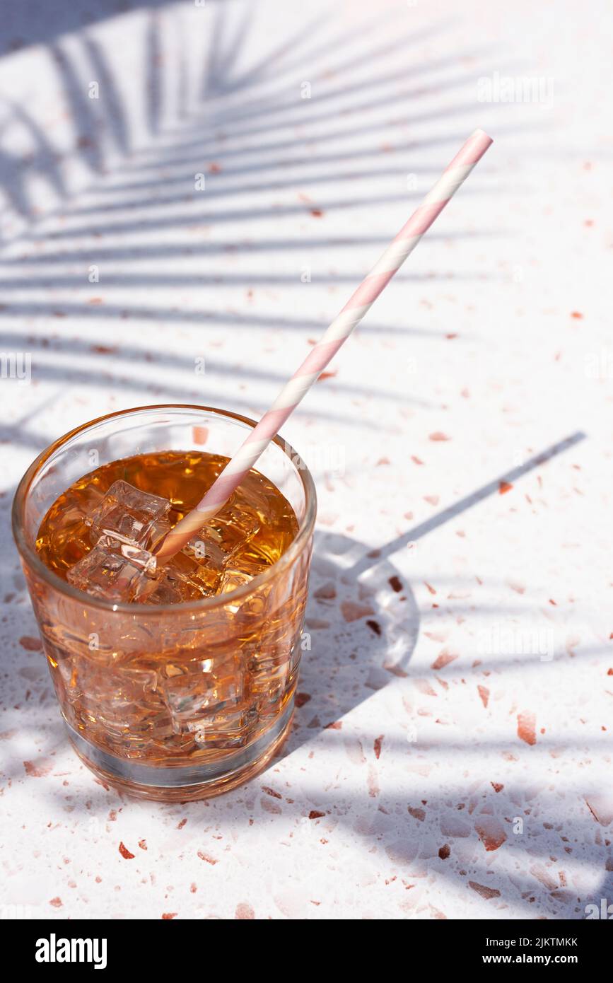A vertical closeup of a glass of juice with ice cubes and straw on a marble table in sunlight Stock Photo