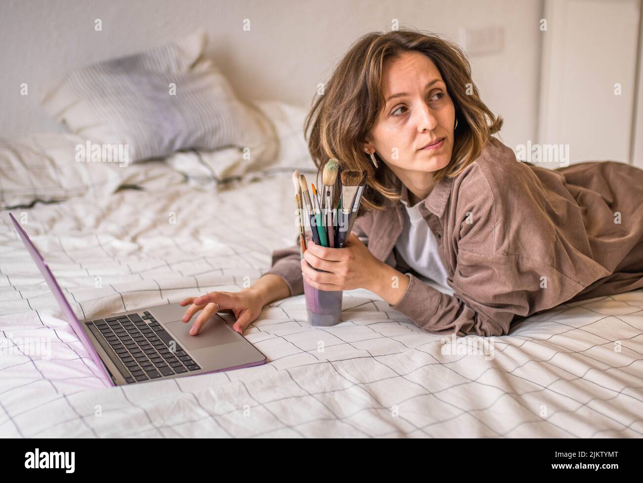 Picture of Thoughtful 30's Slavic Woman Deciding What to Paint Holding a Can of Brushes in One Hand and Typing on her Laptop from her Bedroom Bed Stock Photo