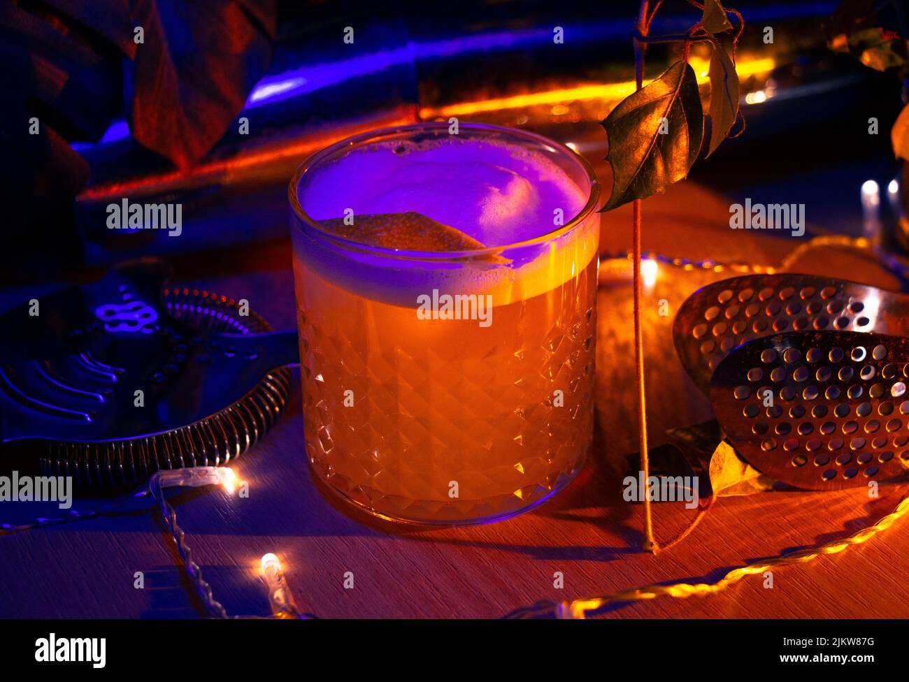 A closeup of a gin fizz cocktail, lemon twist garnish, and bar equipment under neon light Stock Photo