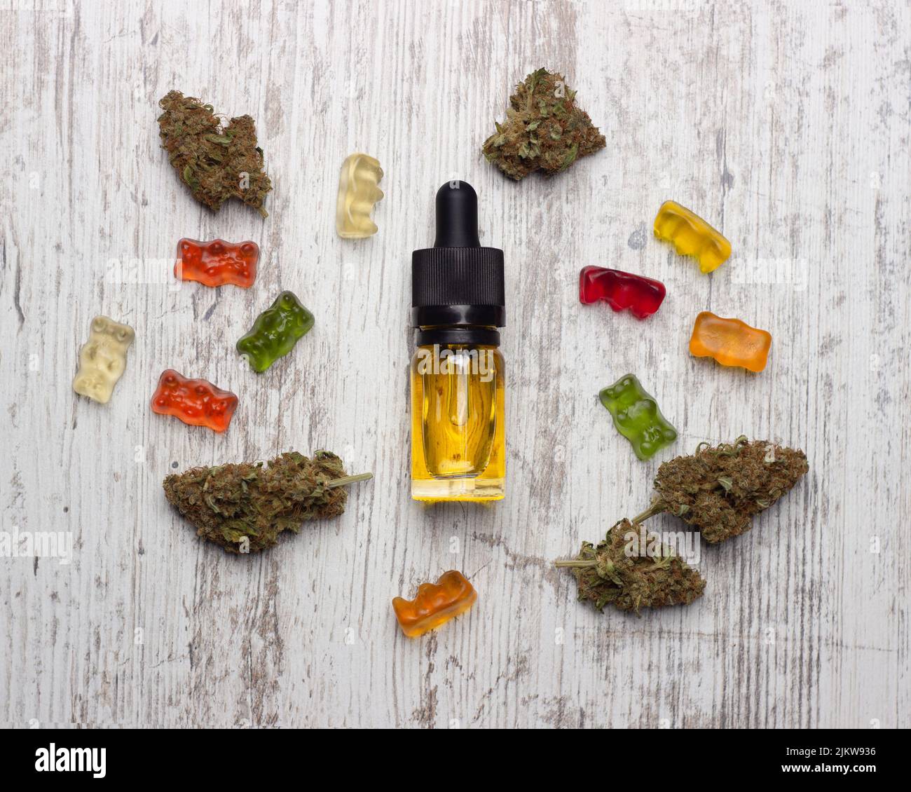 A glass bottle with yellow cannabis cbd oil lies on a wooden table surrounded by dry medical marijuana buds and colorful cbd gummy bears Stock Photo