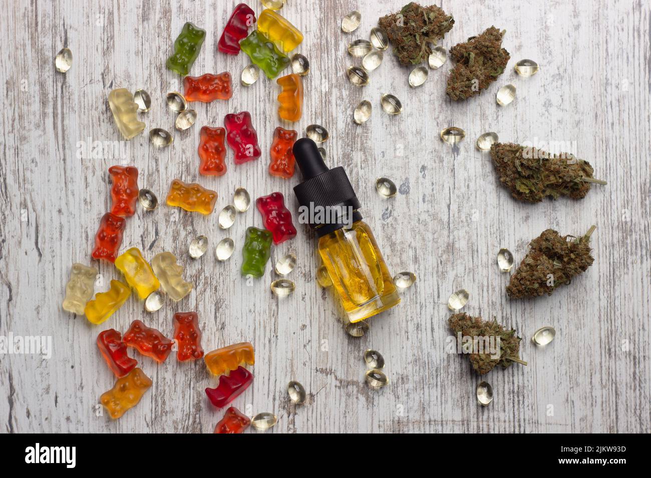 A glass bottle with yellow cannabis cbd oil lies on a wooden table surrounded by dry medical marijuana buds, cbd extract capsules and colorful cbd gum Stock Photo