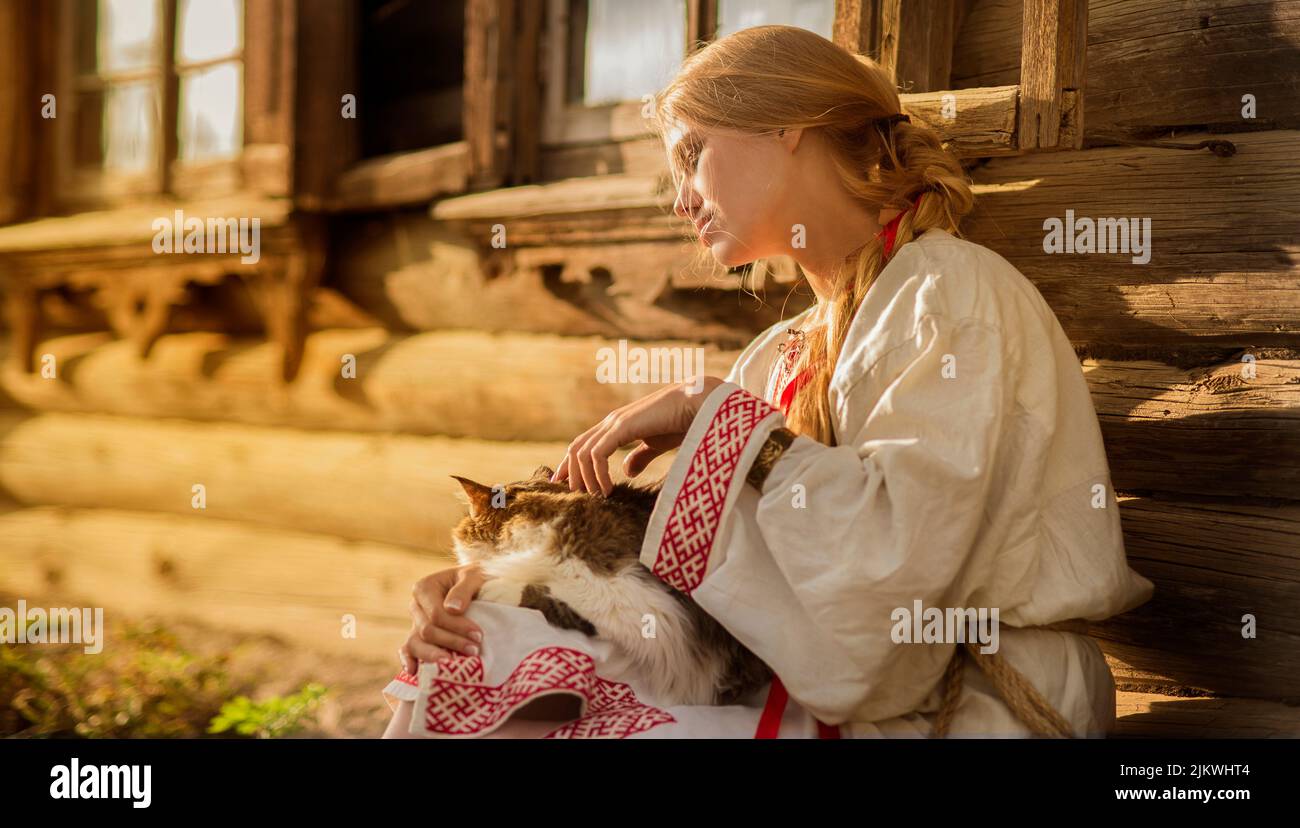 A young woman in a national dress sits near the hut with a cat on her knees during sunset. Russian, Ukrainian or Belarusian national culture. Stock Photo