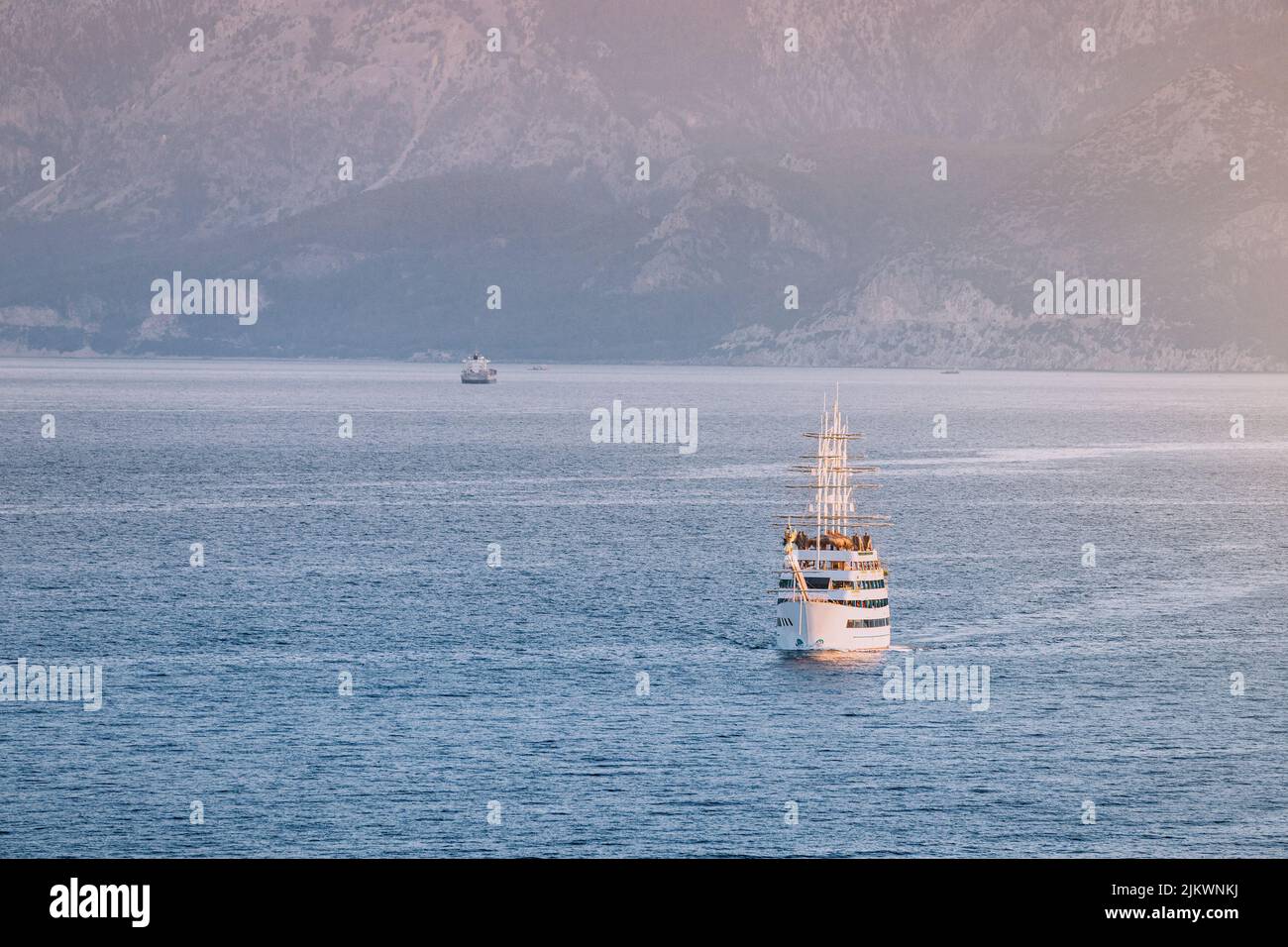 The excursion luxury ship frigate sails on the water surface of the Mediterranean Sea against the backdrop of high mountains and sunset. Stock Photo
