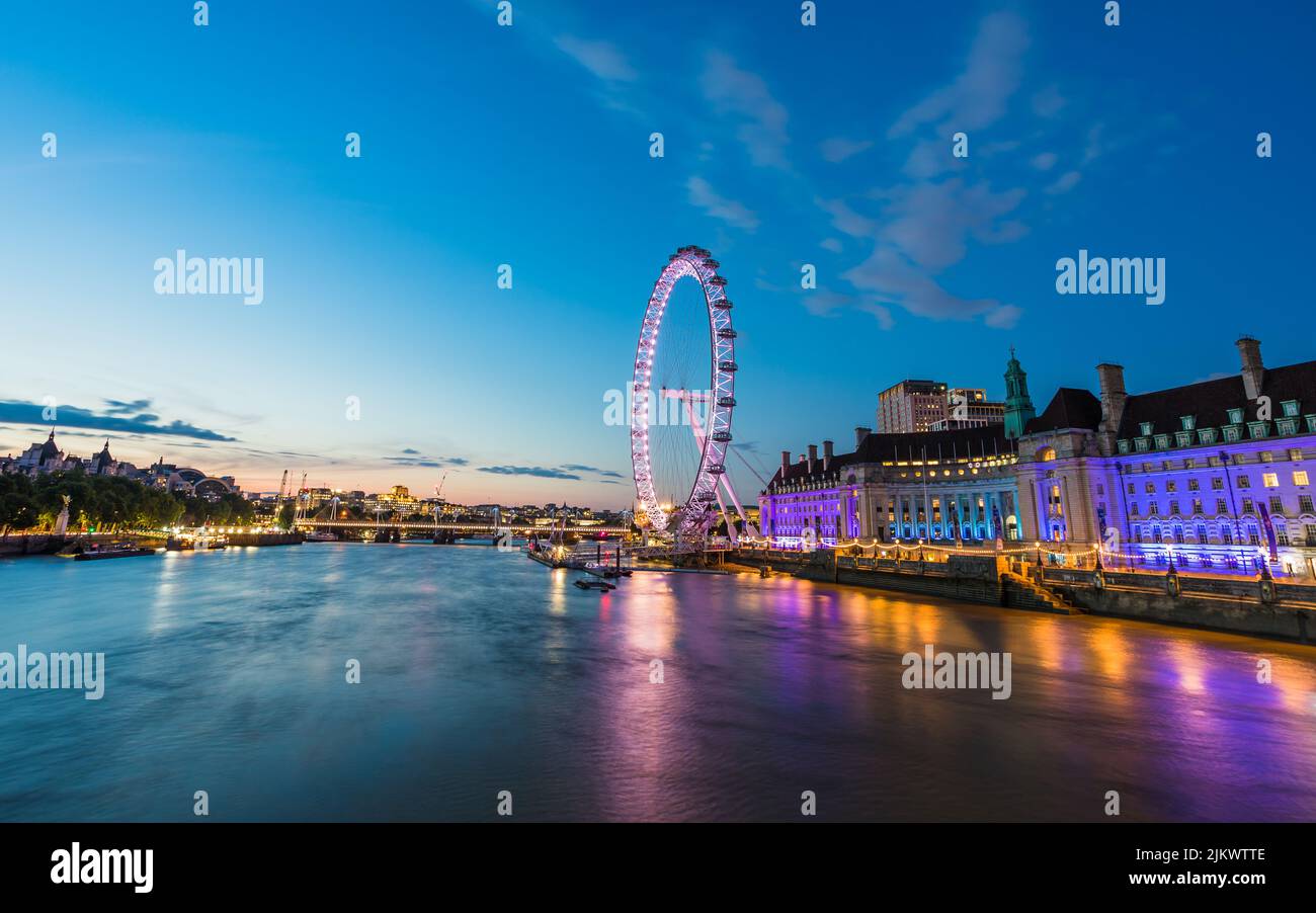 Beautiful tones from the lights on the London Aquarium and the London Eye reflect on the River Thames in London at twilight in July 2022. Stock Photo