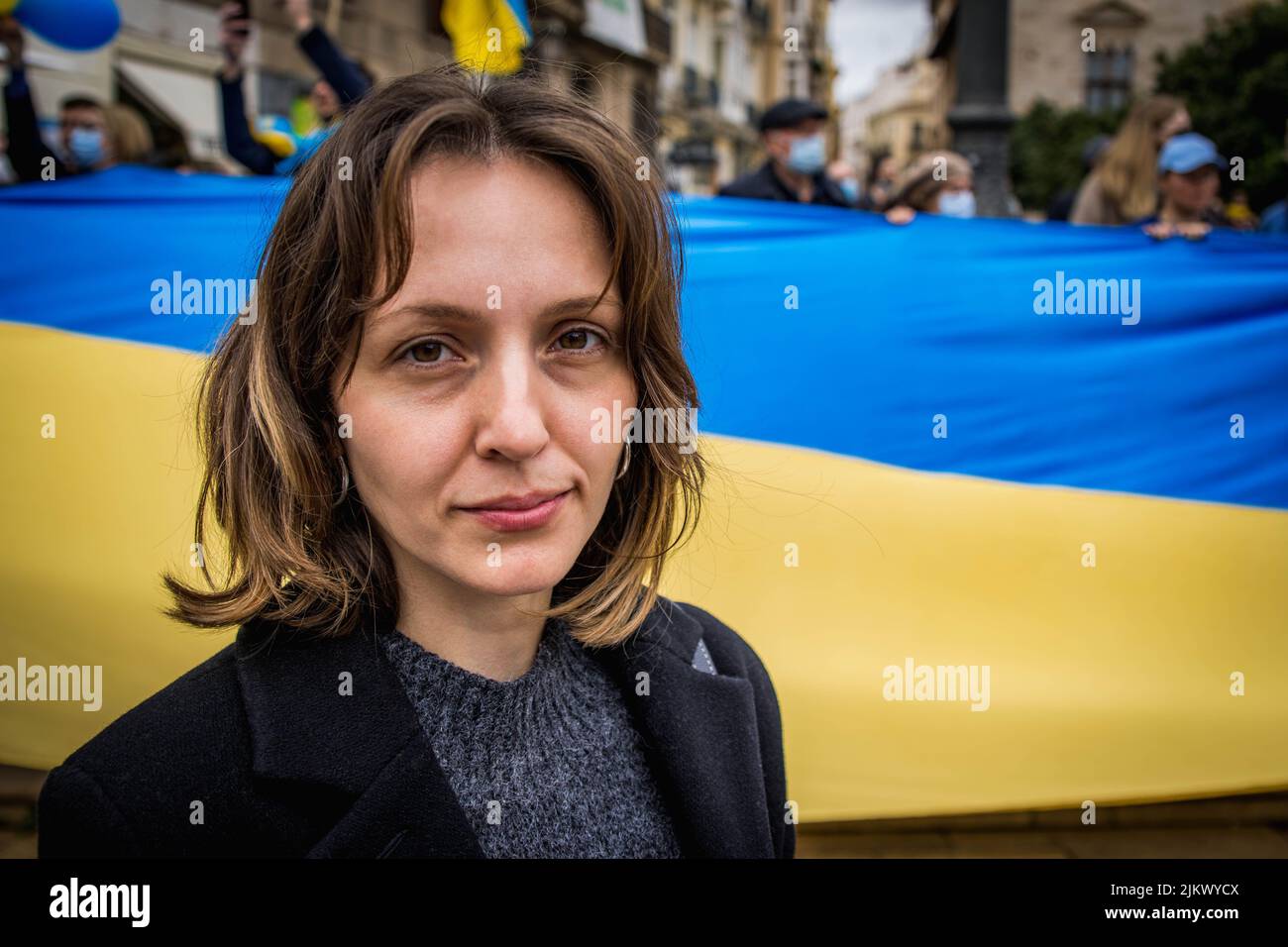 Ukrainian Refugee at Demonstration Against War with Russia Looking Optimistically at Camera with Giant National Flag in Background Stock Photo