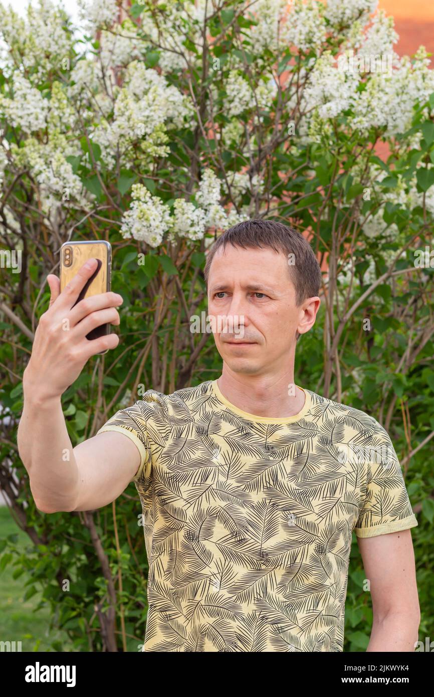 man takes selfie on his cell phone on white lilacs blooming background, close-up, side view, selective focus. Natural background and using technology Stock Photo