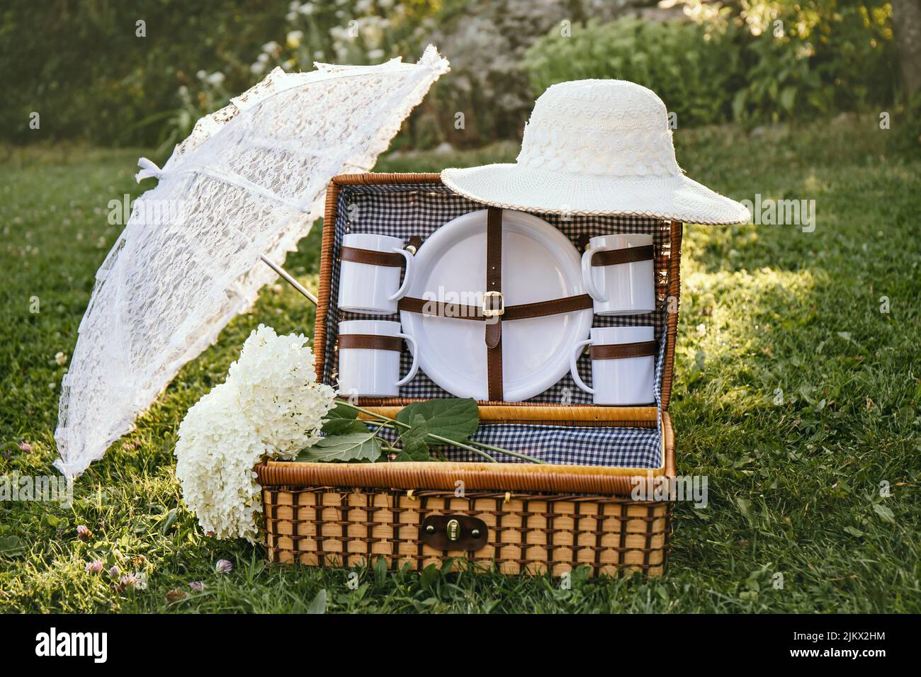 Romantic set with vintage picnic basket, lace sun umbrella and summer hat on park garden lawn on sunny summer day outdoors. Stock Photo