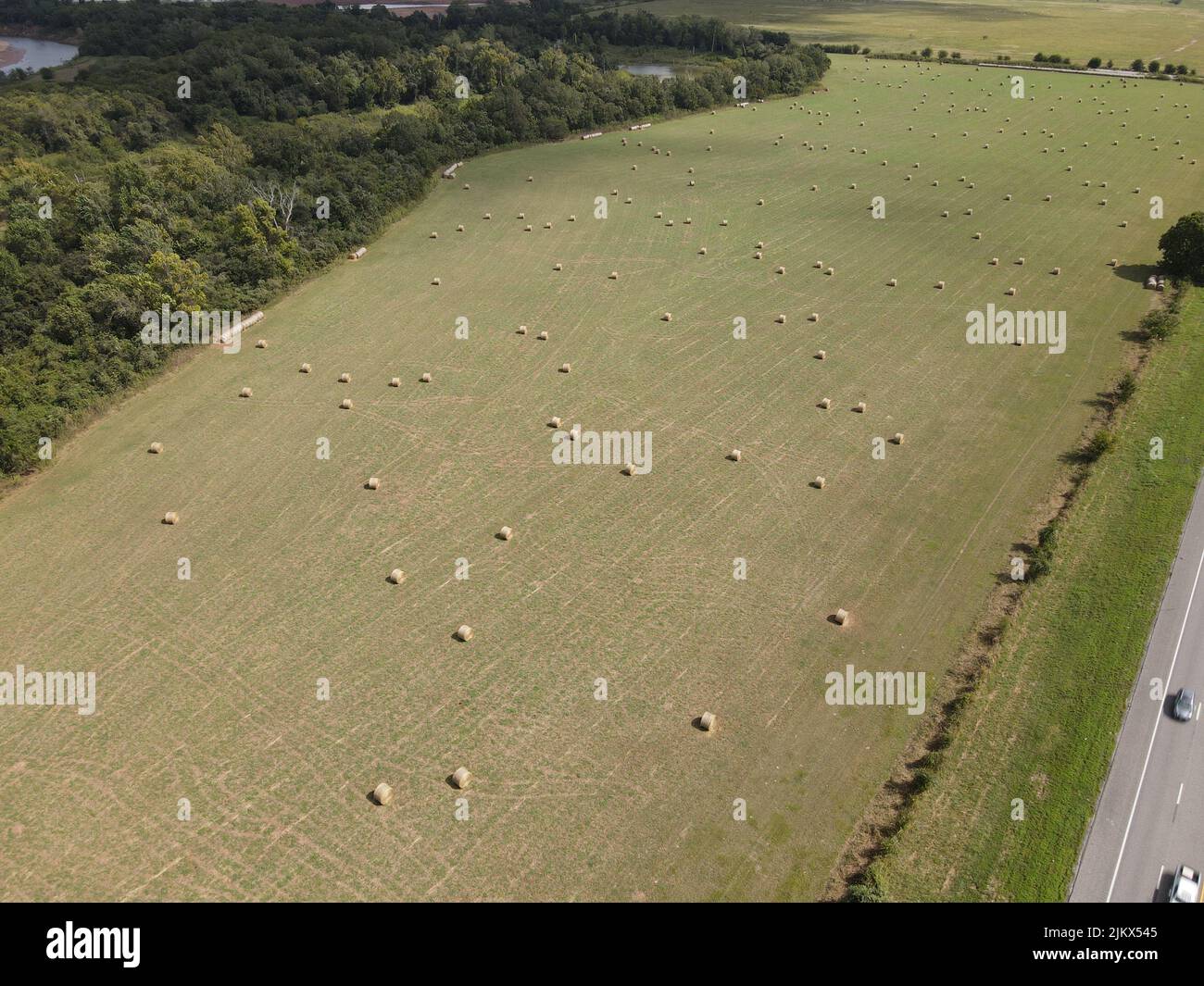 A bird's eye view of hay bales on a green field near cars driving on an asphalt road Stock Photo