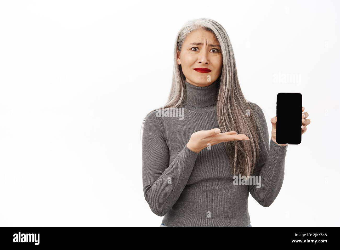 Upset and disappointed asian senior woman, middle aged korean female model pointing, showing smartphone screen and feeling sad, white background Stock Photo