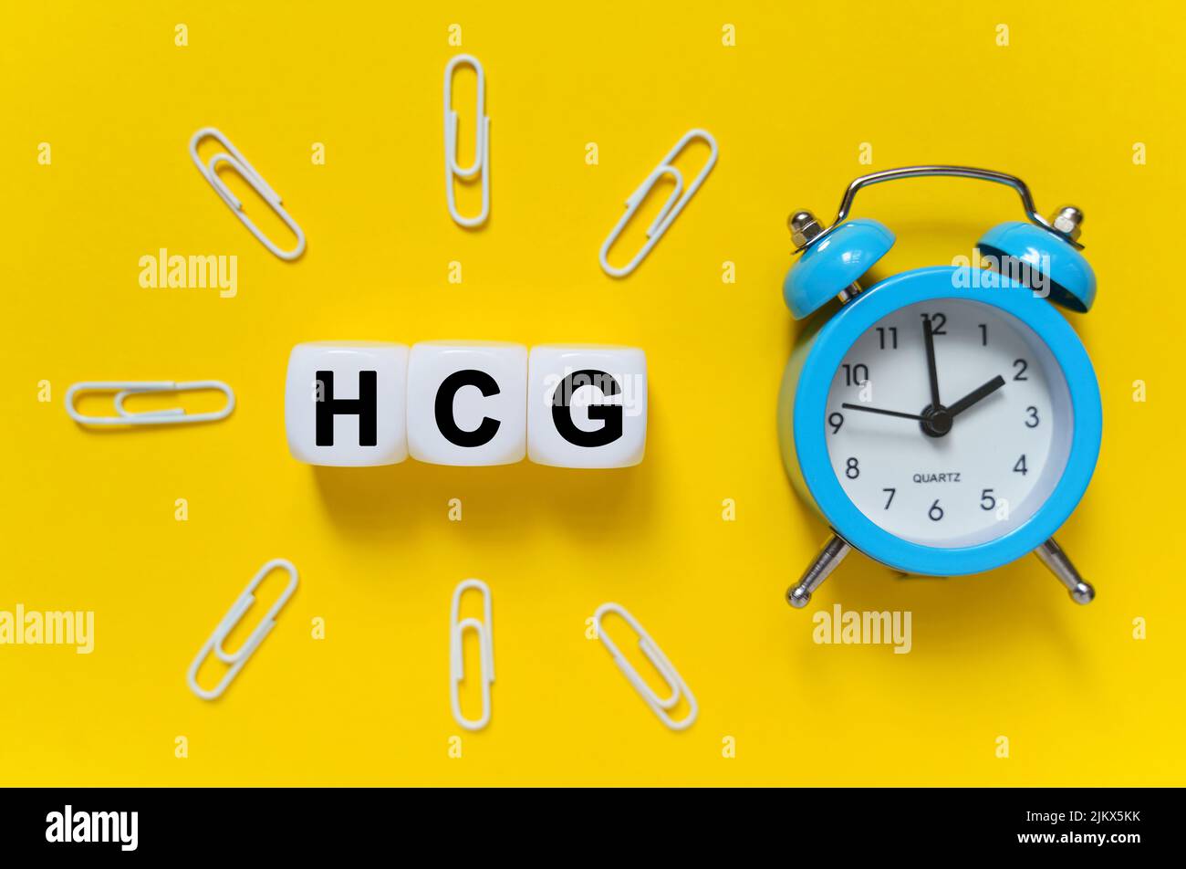 Finance and economics concept. On a yellow background, a blue alarm clock, paper clips and white cubes on which the text is written - HCG Stock Photo