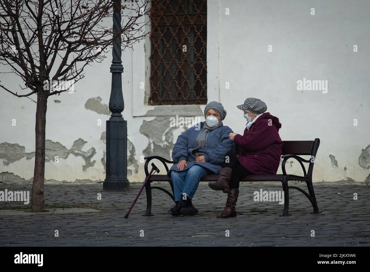 Óbuda, Hungary - Mar 21 2021: Two old ladies sitting on a bench and chatting during Covid time, wearing masks Stock Photo