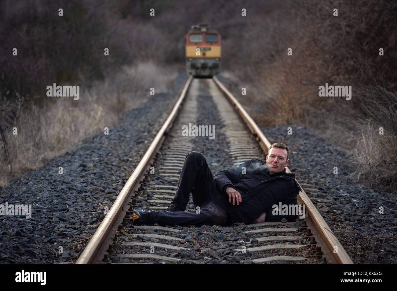 Young man lying on the railway track smoking a cigarette while a train is coming in the background Stock Photo