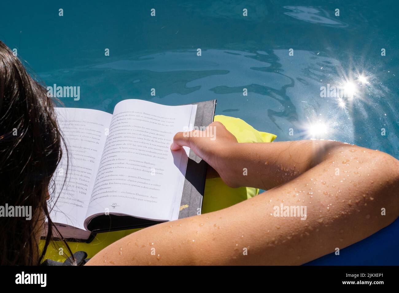 Young girl reading a book while inside a water pool refreshing on summer days. Water sun glare reflexion. Open book, kids reading on hollydays. Stock Photo