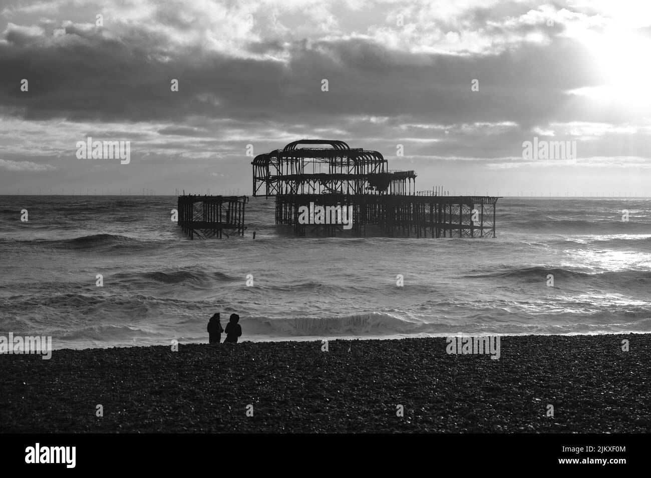 A grayscale view of the seaside in Brighton, an old pier and two people relaxing on the beach Stock Photo