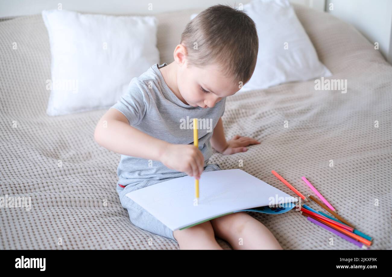 A small caucasian boy in a gray T-shirt and shorts on the bed draws with colored pencils in a sketch Stock Photo