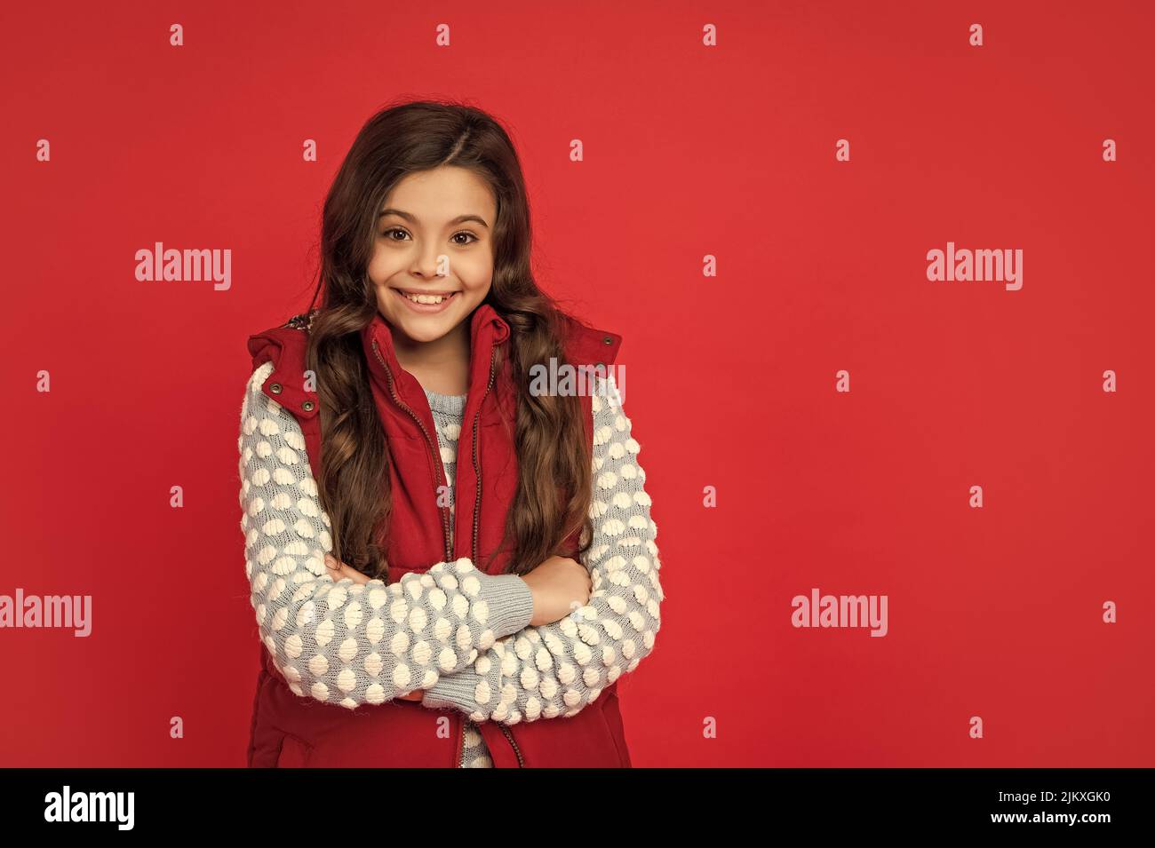 smiling child with curly hair in winter down vest on red background, copy space, wintertime. Stock Photo