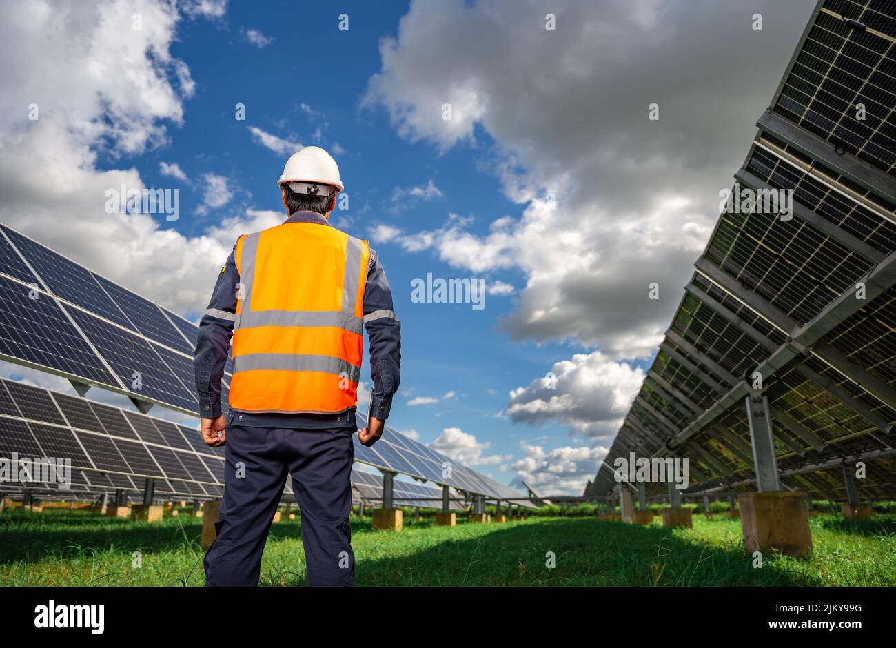 Technician dressed in blue uniform and in an orange reflective vest and wearing a safety helmet stands facing a solar panel at solar power plant, Phot Stock Photo