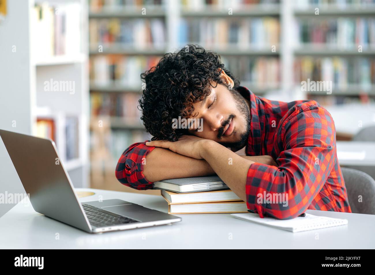 Tired bored arabian or indian university student, is sleeping while sitting at a desk in a library, exhausted of learning, preparing for exam session, suffering of stress and chronic sleep deprivation Stock Photo
