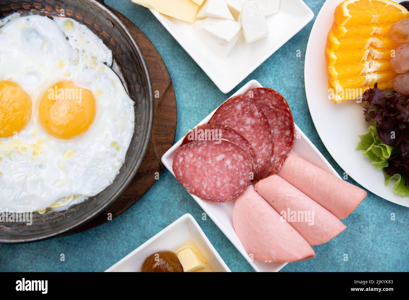 A top view of a breakfast table with sunny side up eggs in a pan and cheeses, cured meats, and fruits Stock Photo