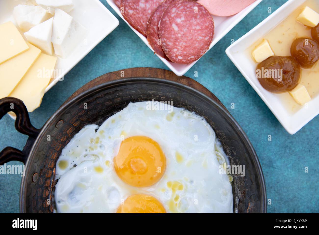 A top view of a breakfast table with sunny side up eggs in a pan and cheeses, cured meats, and jam Stock Photo