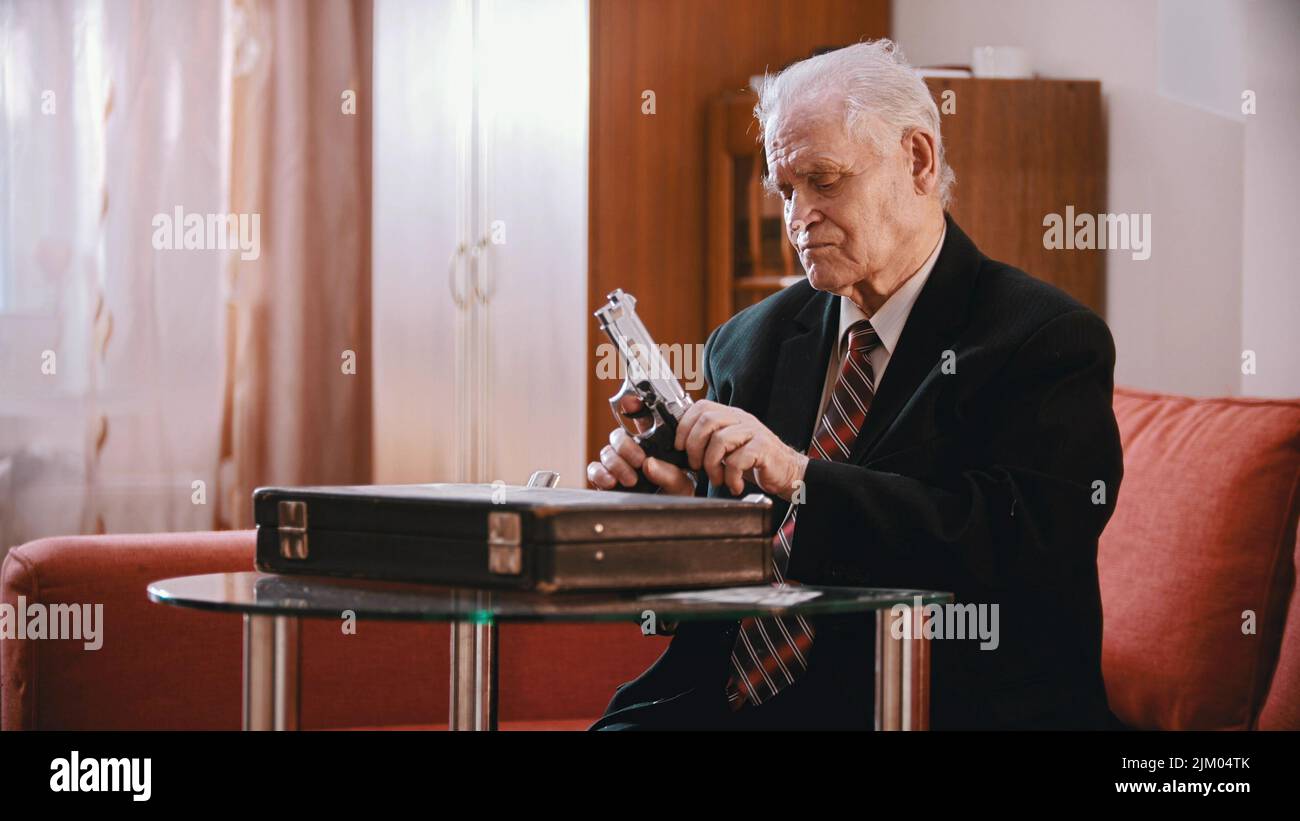 An old sad businessman sitting by the suitcase and reloading the gun ...