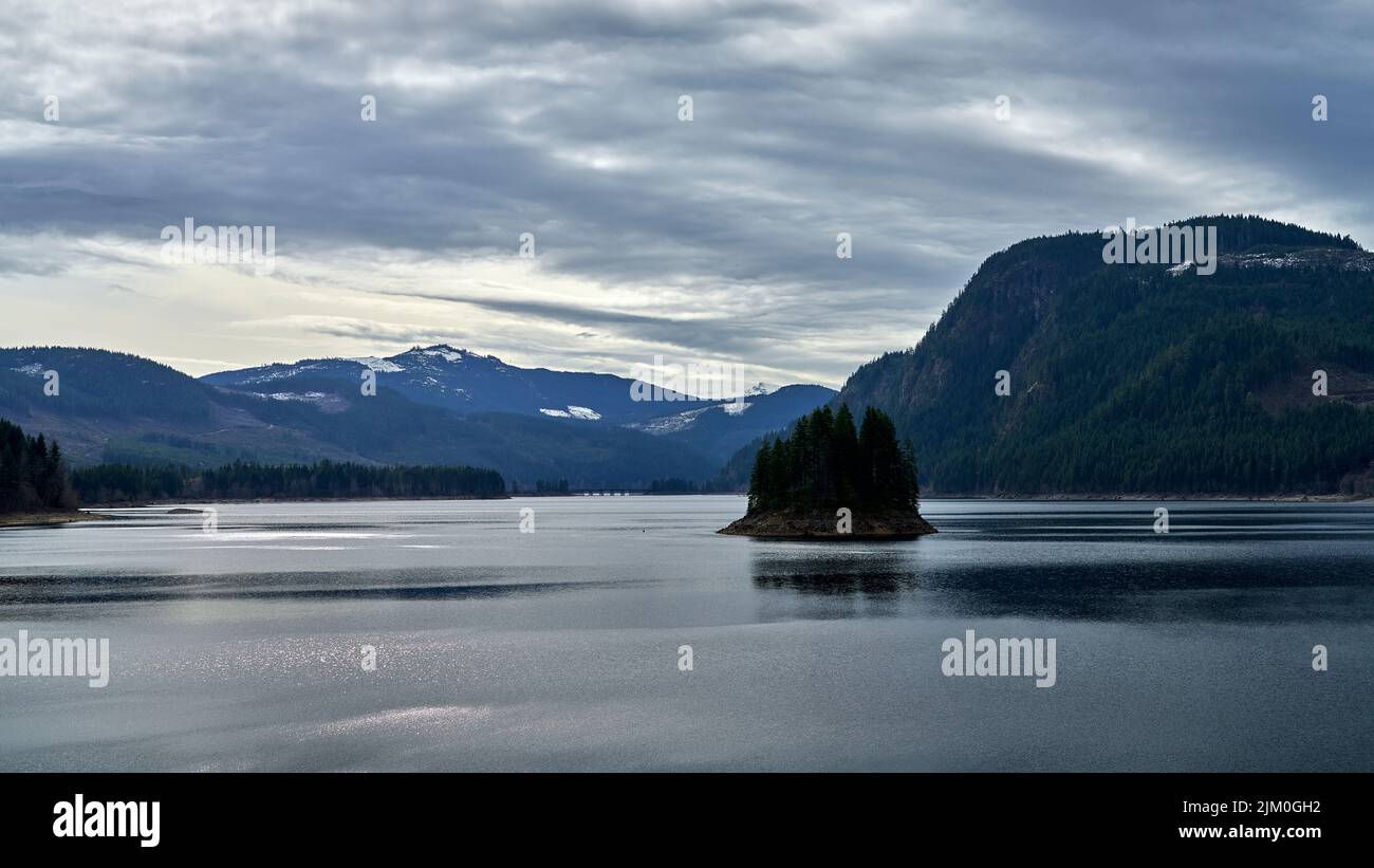 View of a beautiful calm lake, the distant snow capped mountains and a dramatic sky. Stock Photo