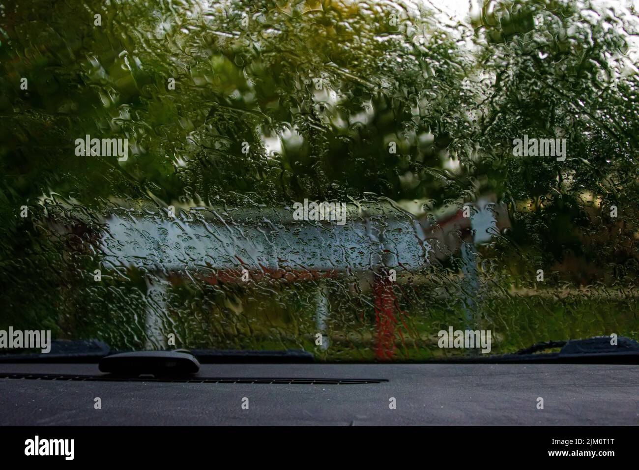 A closeup of a rainy car windshield looking out into a park with a Bluetooth earpiece on the dashboard Stock Photo