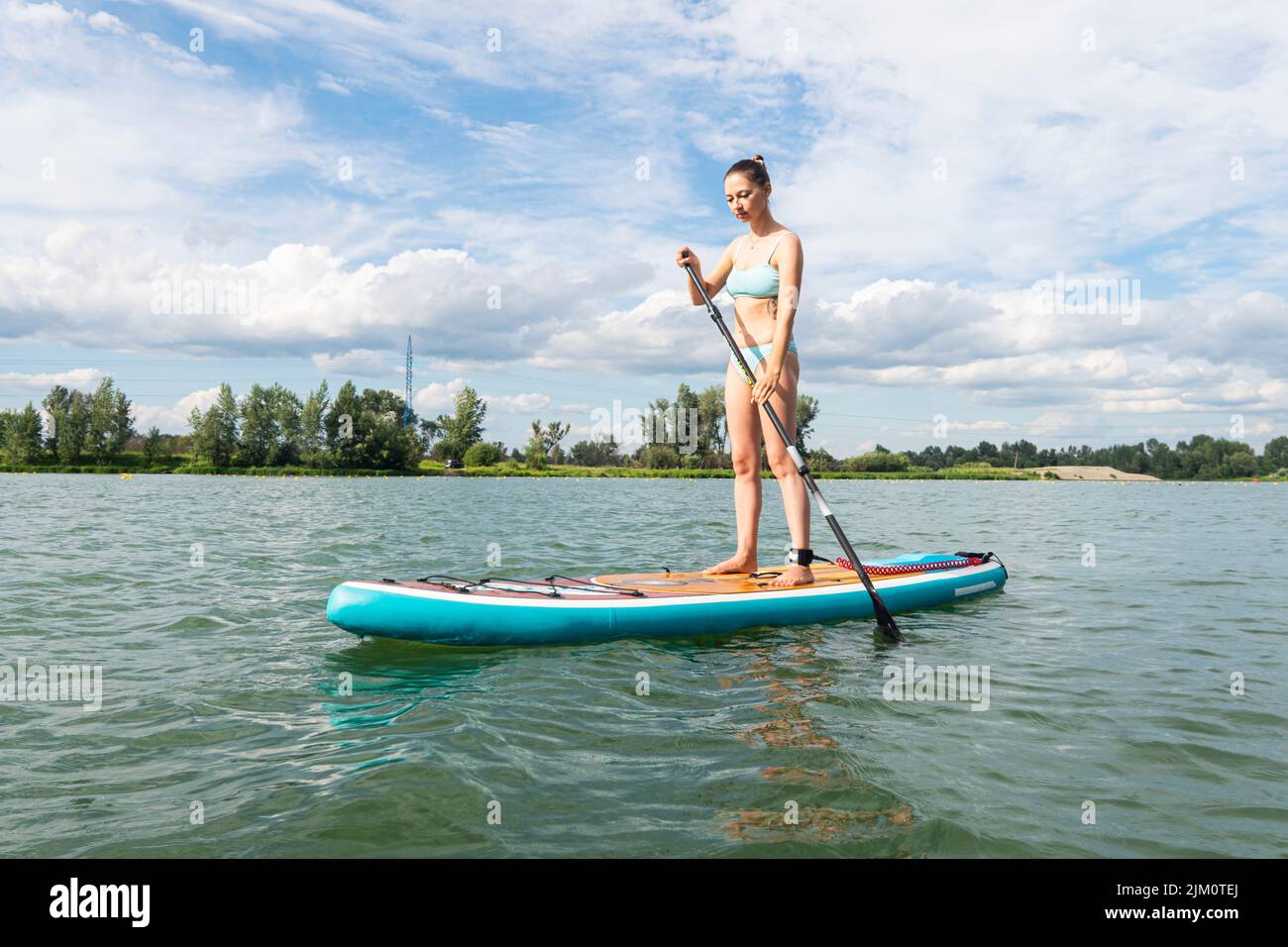 Caucasian woman standing on sup board with one oar in hands at sky on crystal blue lake wearing summer blue swimsuit dress. Active lifestyle. Stock Photo