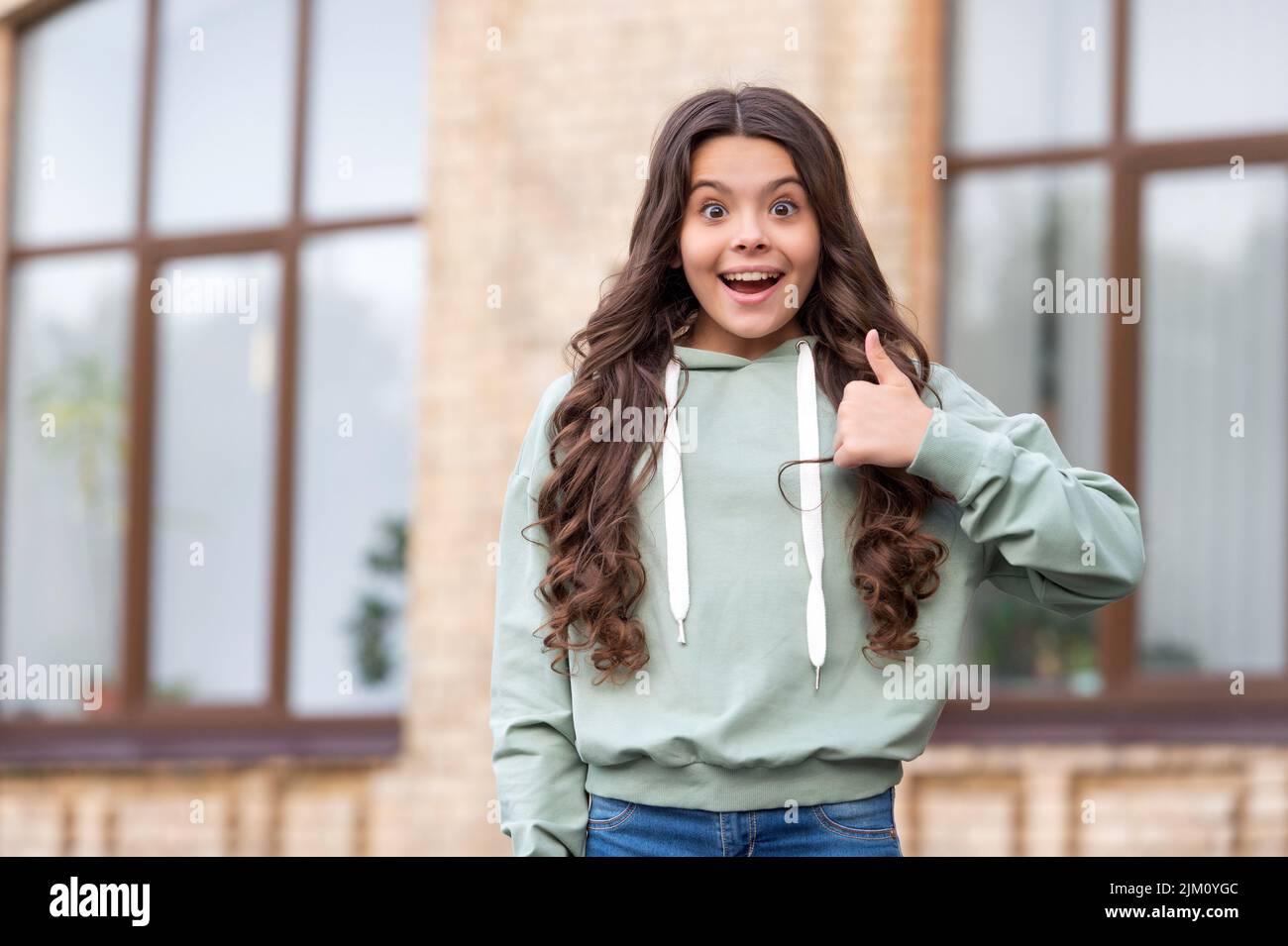 Surprised teenage girl in casual hoodie giving thumb gesture blurry outdoors, copy space Stock Photo