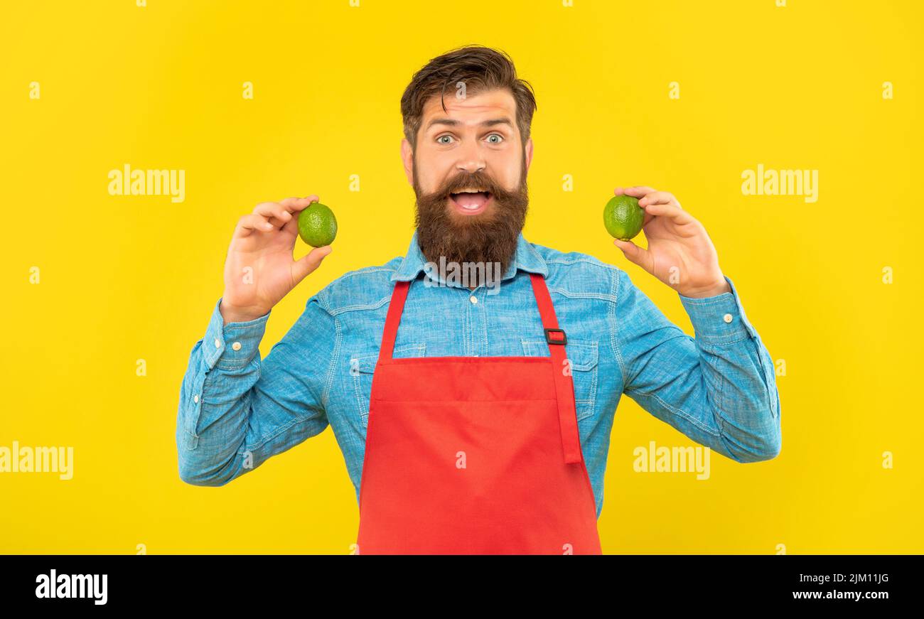 Surprised man in red apron holding fresh limes citrus fruits yellow background, fruiterer Stock Photo