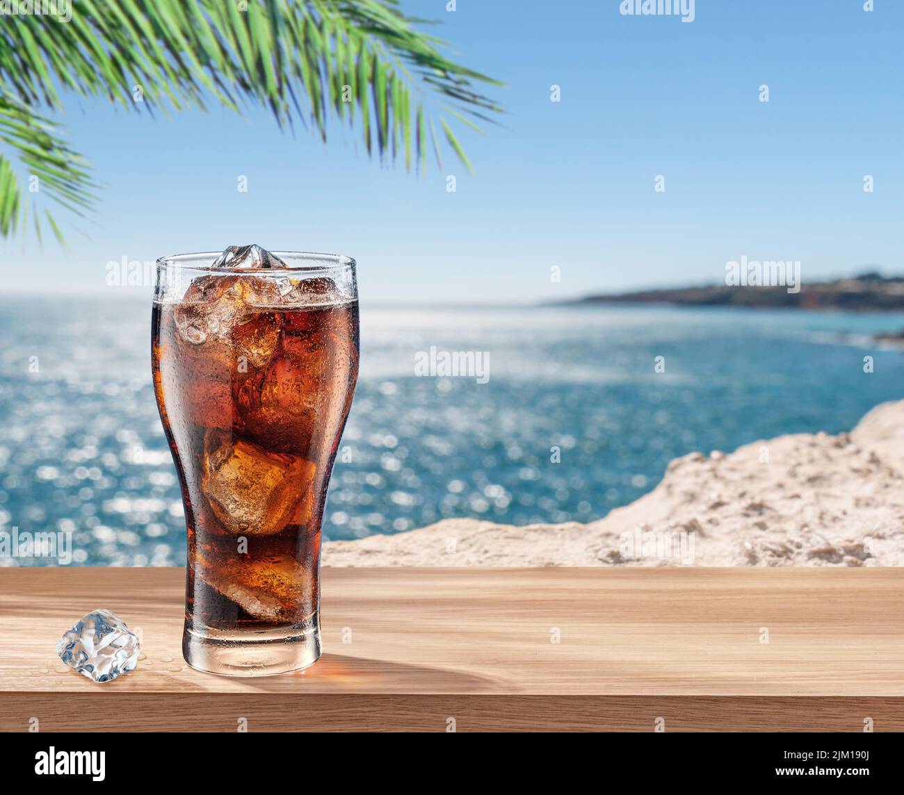 Glass of cola drink with ice cubes on table and blurred sparkling sea at the background. Stock Photo