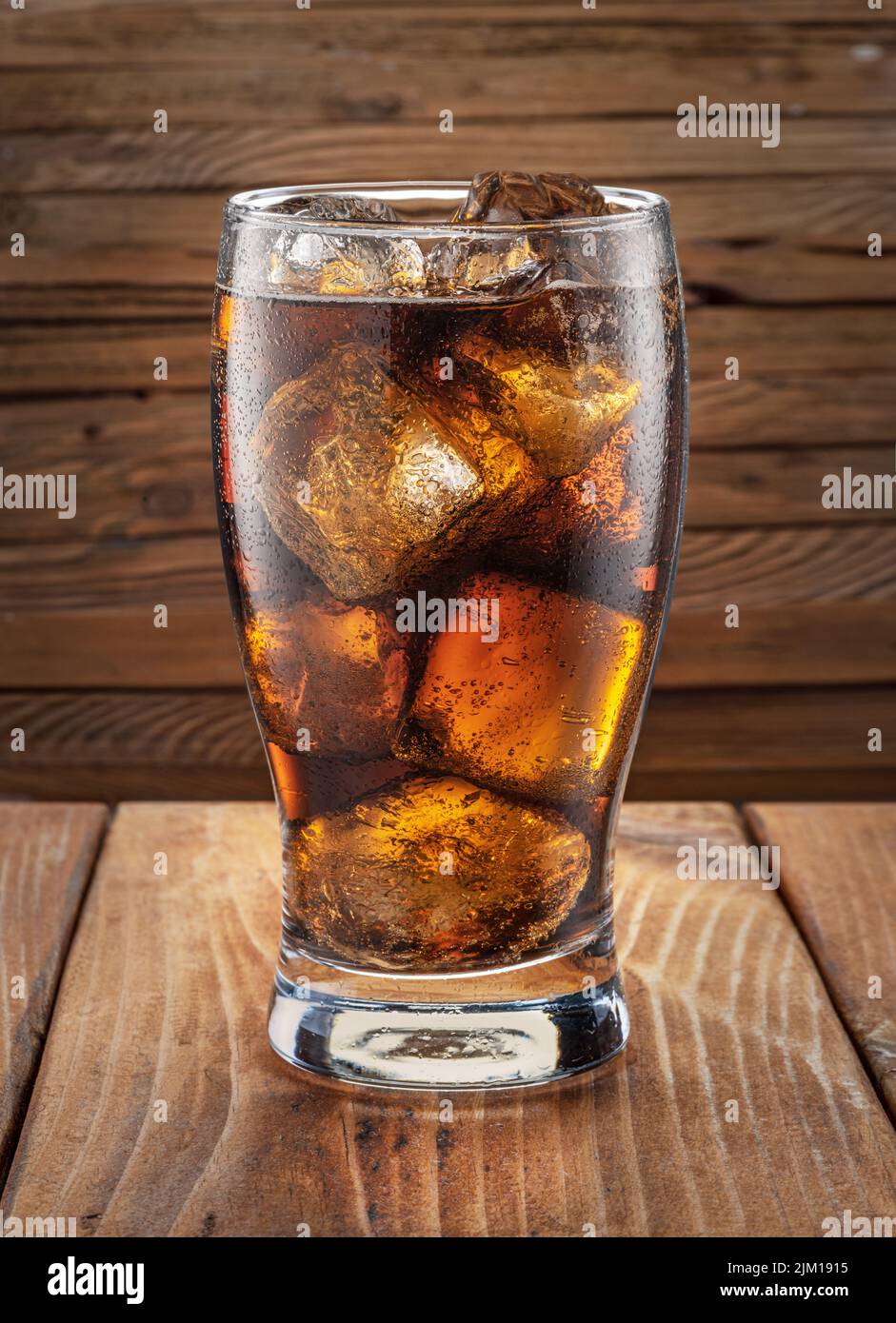 Glass of cola drink with ice cubes on plank table and plank wooden wall at the background. Stock Photo