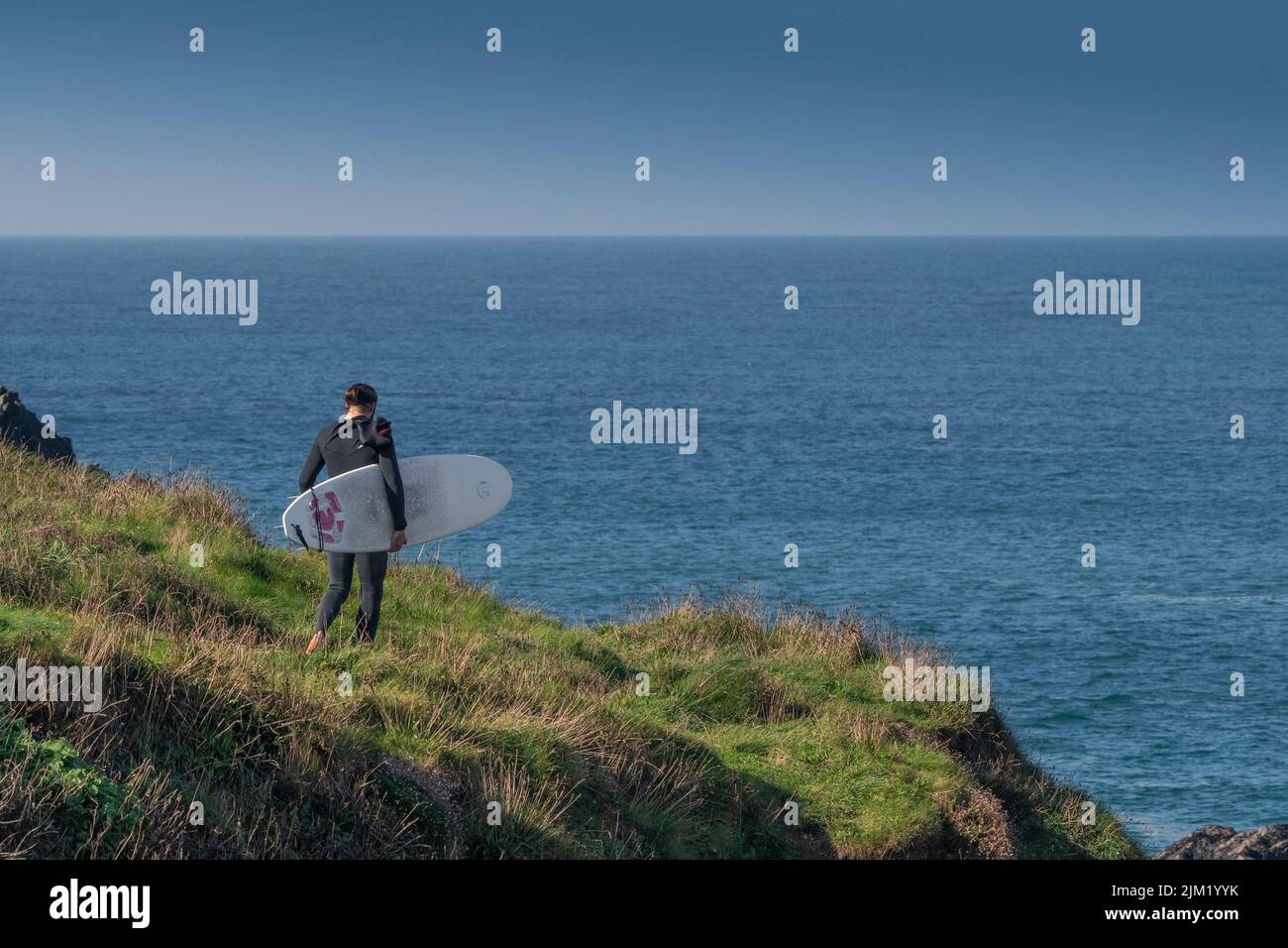 A surfer carrying his surfboard walking along a coast path to the sea at Newquay in Cornwall. Stock Photo