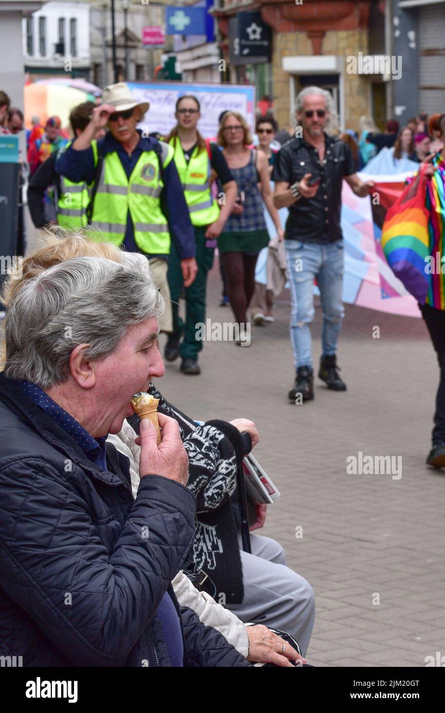 A spectator sitting on a bench and eating an ice cream as the vibrant colourful Cornwall Prides Pride parade passes in Newquay Town centre in the UK. Stock Photo