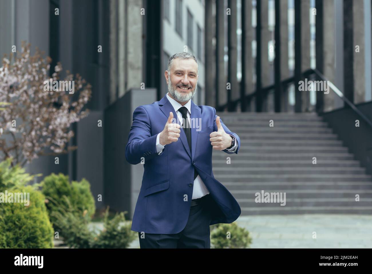 Successful senior gray haired businessman boss, outside office building smiling and looking at camera, experienced investor showing middle finger up, man in business suit Stock Photo