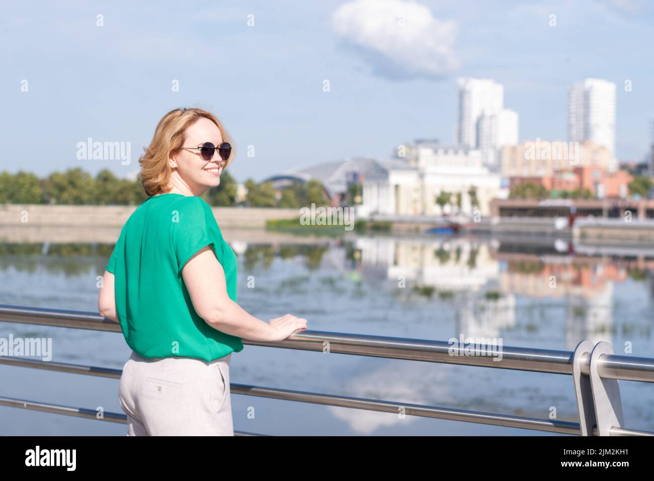 A view from the back of a girl in a green blouse standing on the riverbank against the background of a blurred city.   A girl walks through the street Stock Photo
