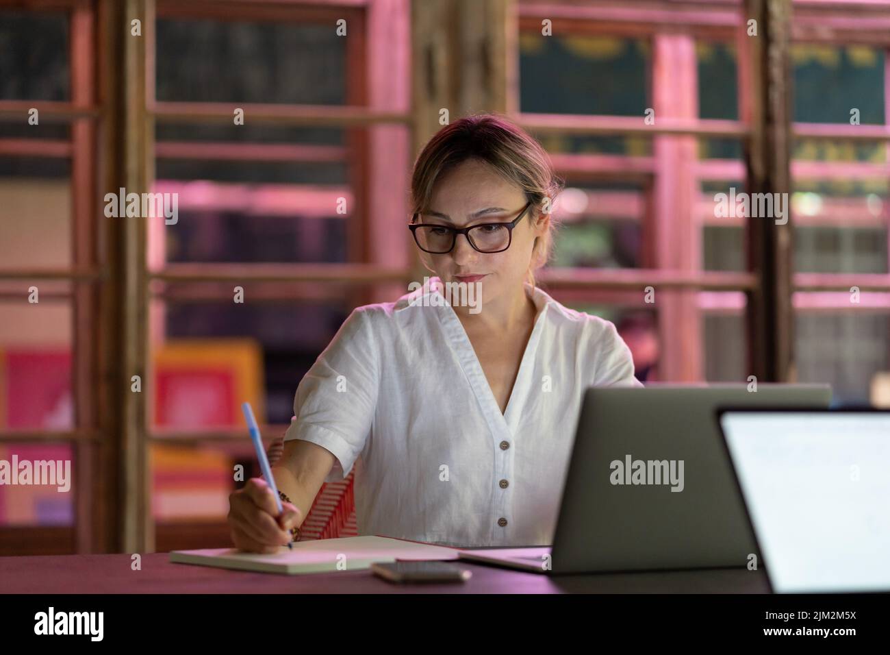 Focused woman wearing glasses watching online course or educational webinar on laptop, making notes Stock Photo