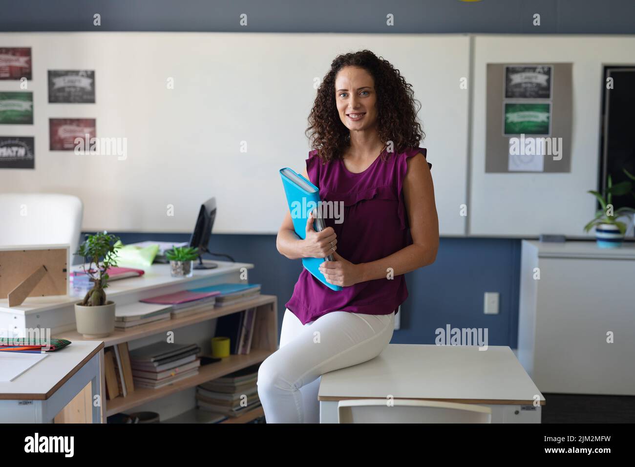 Portrait of smiling caucasian young female teacher with folder sitting on table in staffroom Stock Photo