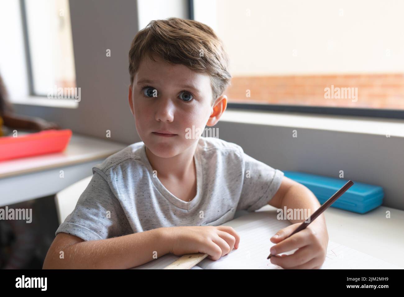 Portrait of caucasian elementary schoolboy with wide eyes sitting at desk in classroom. unaltered, education, childhood, learning and school concept. Stock Photo