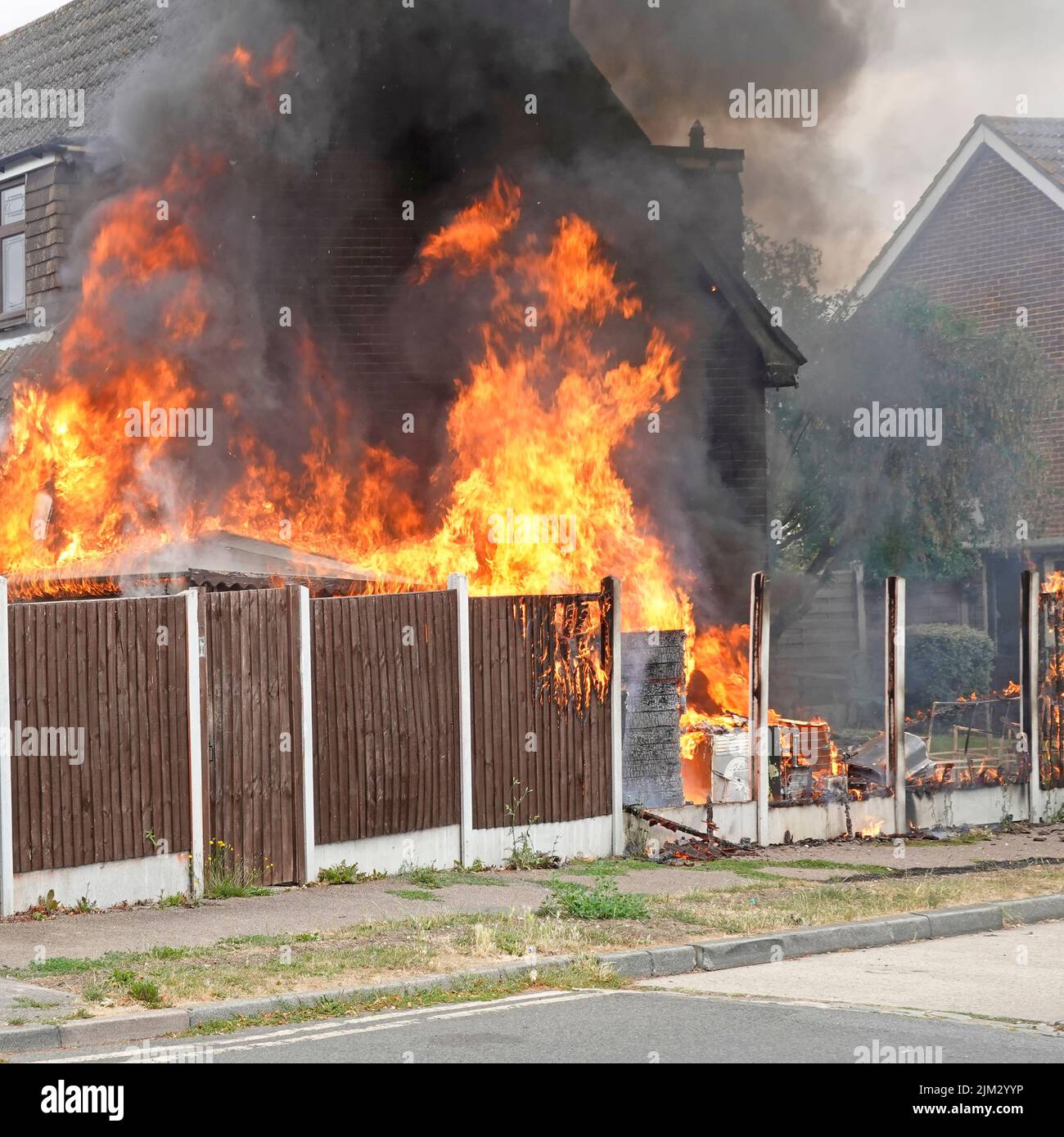 Flames from assorted household items stored outdoors beside timber boundary fence ignited in scorching summer heat setting fire against house wall UK Stock Photo