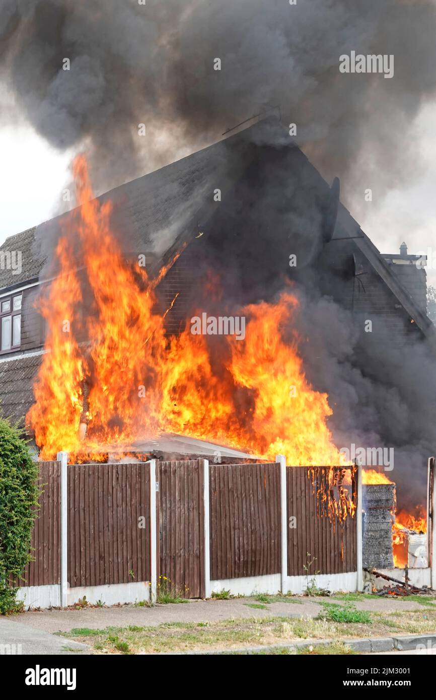 Flames from assorted household items stored outdoors beside timber boundary fence ignited in scorching summer heat setting fire against house wall UK Stock Photo