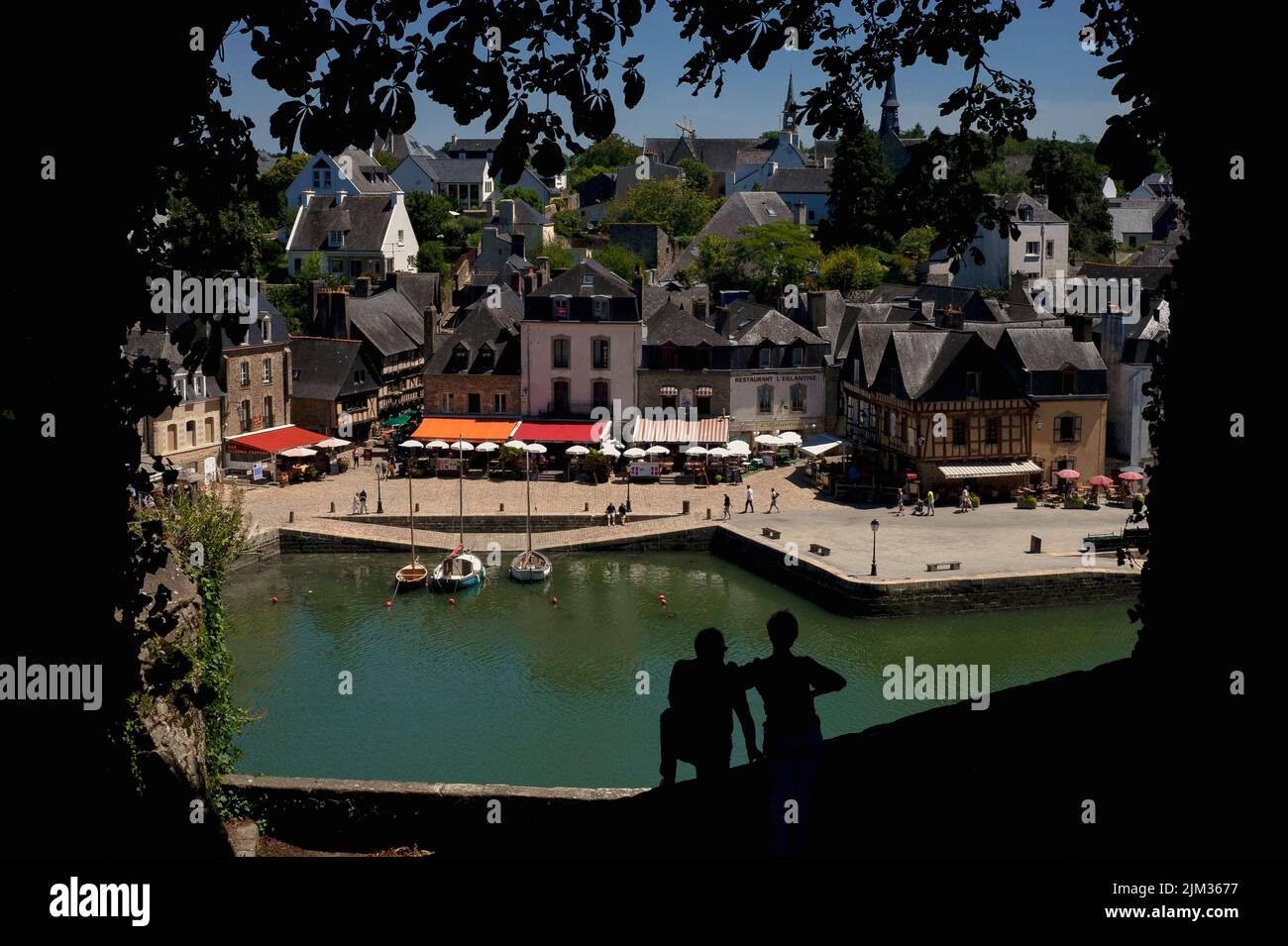 A couple silhouetted against the green River Loch stand looking down on the Place Saint-Sauveur in the Saint-Goustan district of Auray, Morbihan, southern Brittany, France, lined with restaurants and pavement cafes with colourful awnings and sun umbrellas.  Saint-Goustan, once a busy fishing and commercial port, is now a popular tourist destination that honours a 1776 visit by US Founding Father, Benjamin Franklin. Stock Photo