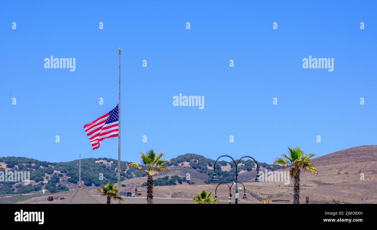 US flag at half-mast at Pismo Beach, Central California, USA. Blue sky & hills in background. Stock Photo