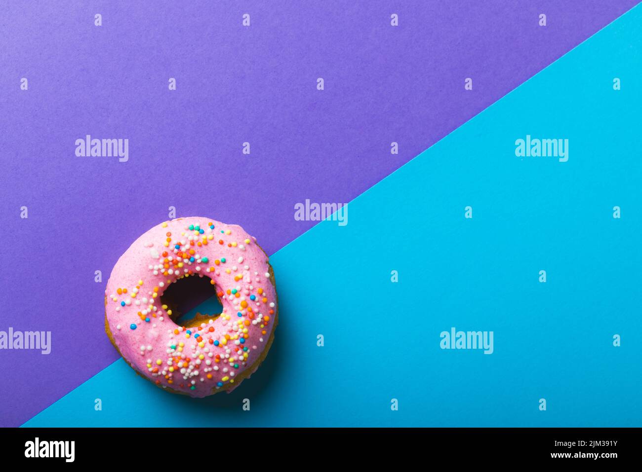Directly above view of fresh pink donut with sprinklers by copy space against two tone background. unaltered, unhealthy eating and sweet food concept. Stock Photo