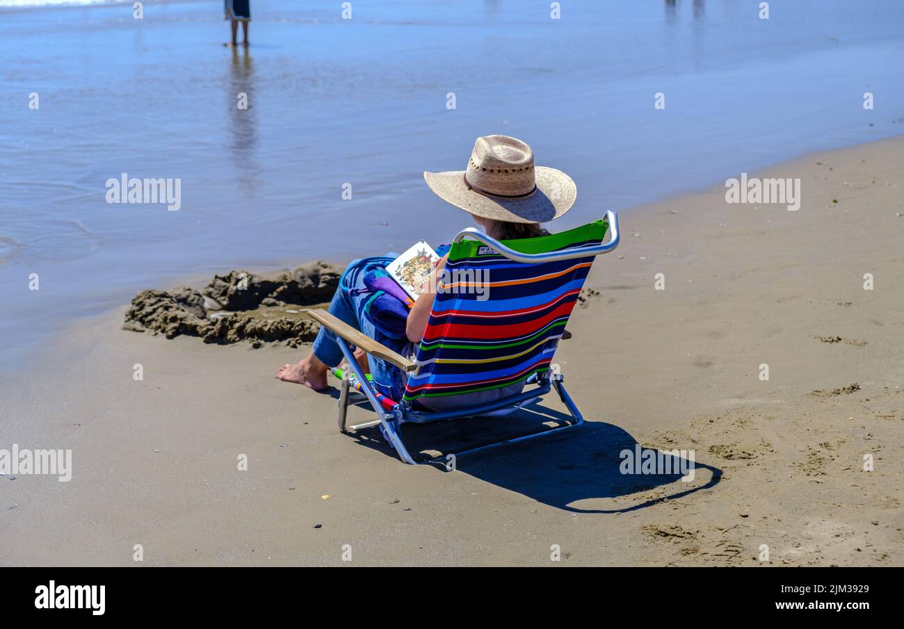 Lady in sun hat sits in chair at water’s edge reading a book at Pismo Beach,  Central California. Stock Photo
