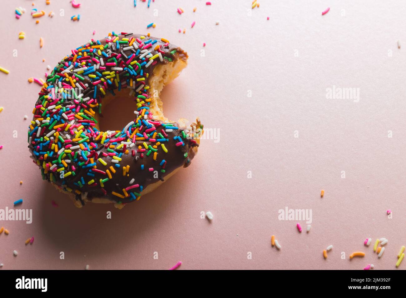 Overhead view of missing bite of chocolate donut with sprinklers by copy space on pink background. unaltered, unhealthy eating and sweet food concept. Stock Photo