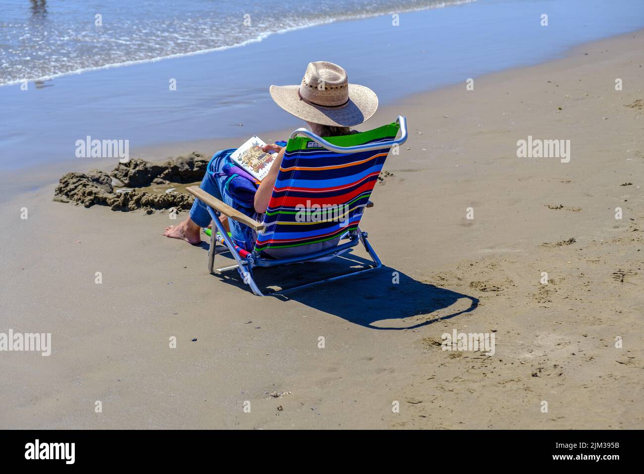 Lady in sun hat sits in chair at shoreline, reading a book at Pismo Beach, Central California. Stock Photo
